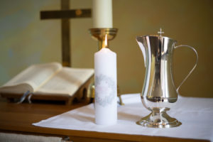 Auf dem Altar ist eine brennende Taufkerze neben einer silbernen Taufkanne zu sehen. Im Hintergrund liegt eine aufgeschlagene Bibel und es ist das Kreuz zu sehen.